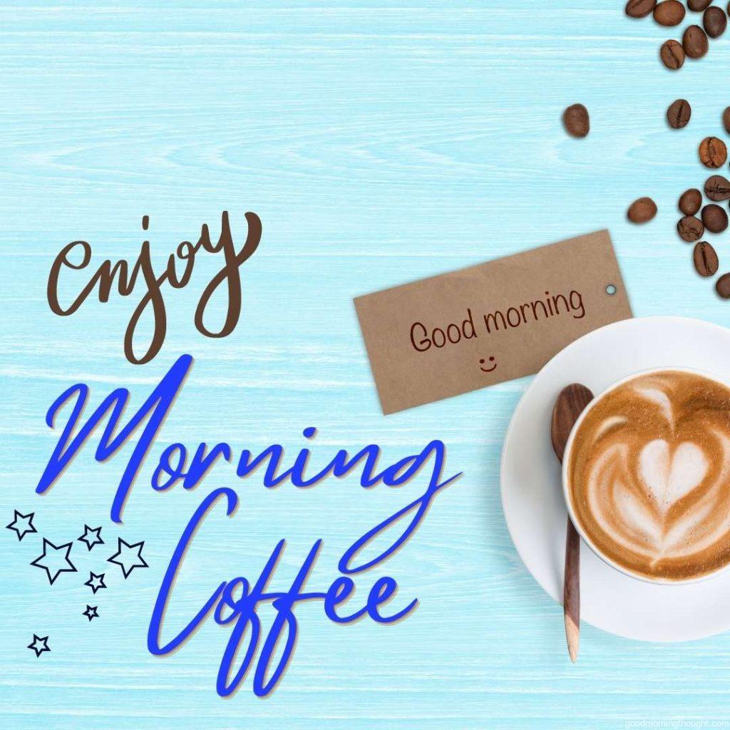 A cup of latte coffee with good morning message smiley and scattered roasted coffee beans on pastel blue desk