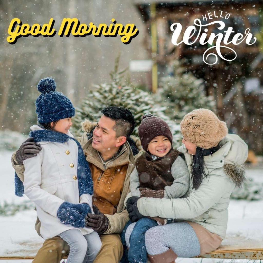 An Asian family is outdoors at a Christmas tree farm during the winter. The father, daughter, mother, and son are sitting and smiling at each other. Snow is falling gently. _Good morning_ text