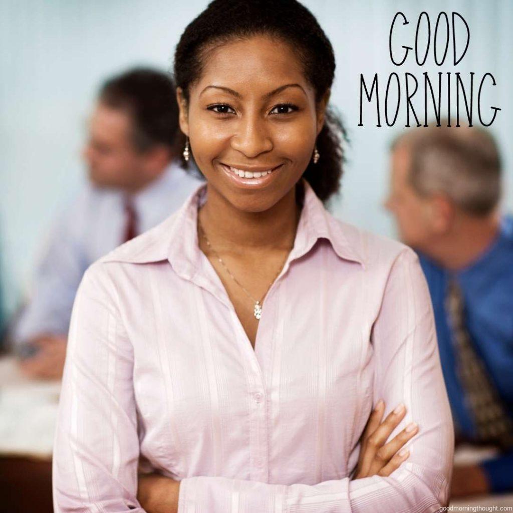 A portrait of an African-American businesswoman in an office with computers that are out of focus in the background A woman is looking at the camera with a toothy smile. Good Morning Text