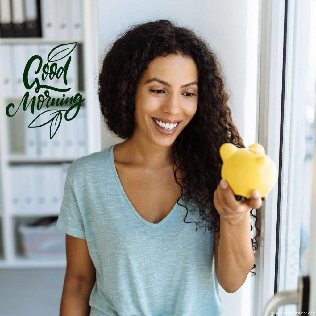 A young African American woman holding a yellow piggy bank with a smile on her face stands in an office environment. Good Morning Text