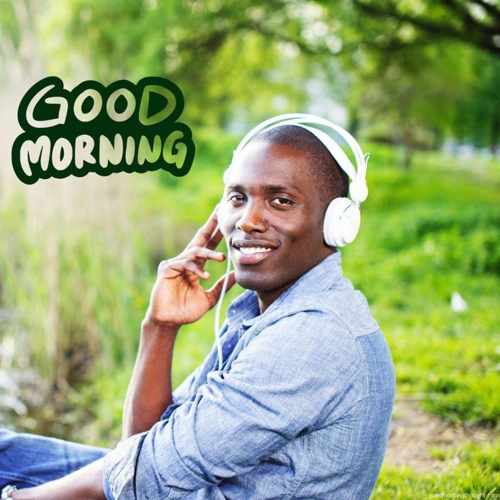 A young, happy, smiling African American sits on grass and listens to music in a park. Good Morning Text