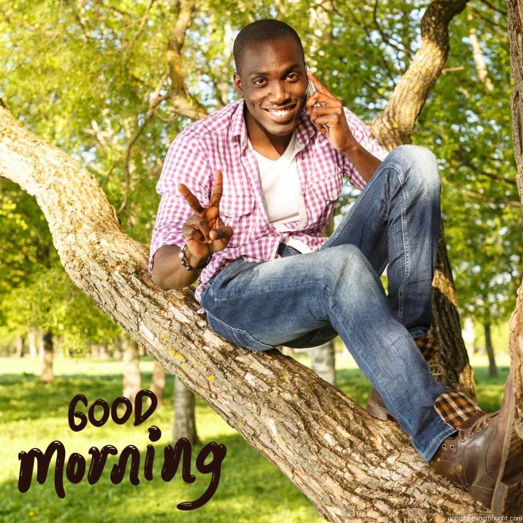 A young, smiling African American sits on a tree in a park with a mobile phone. Good Morning Text