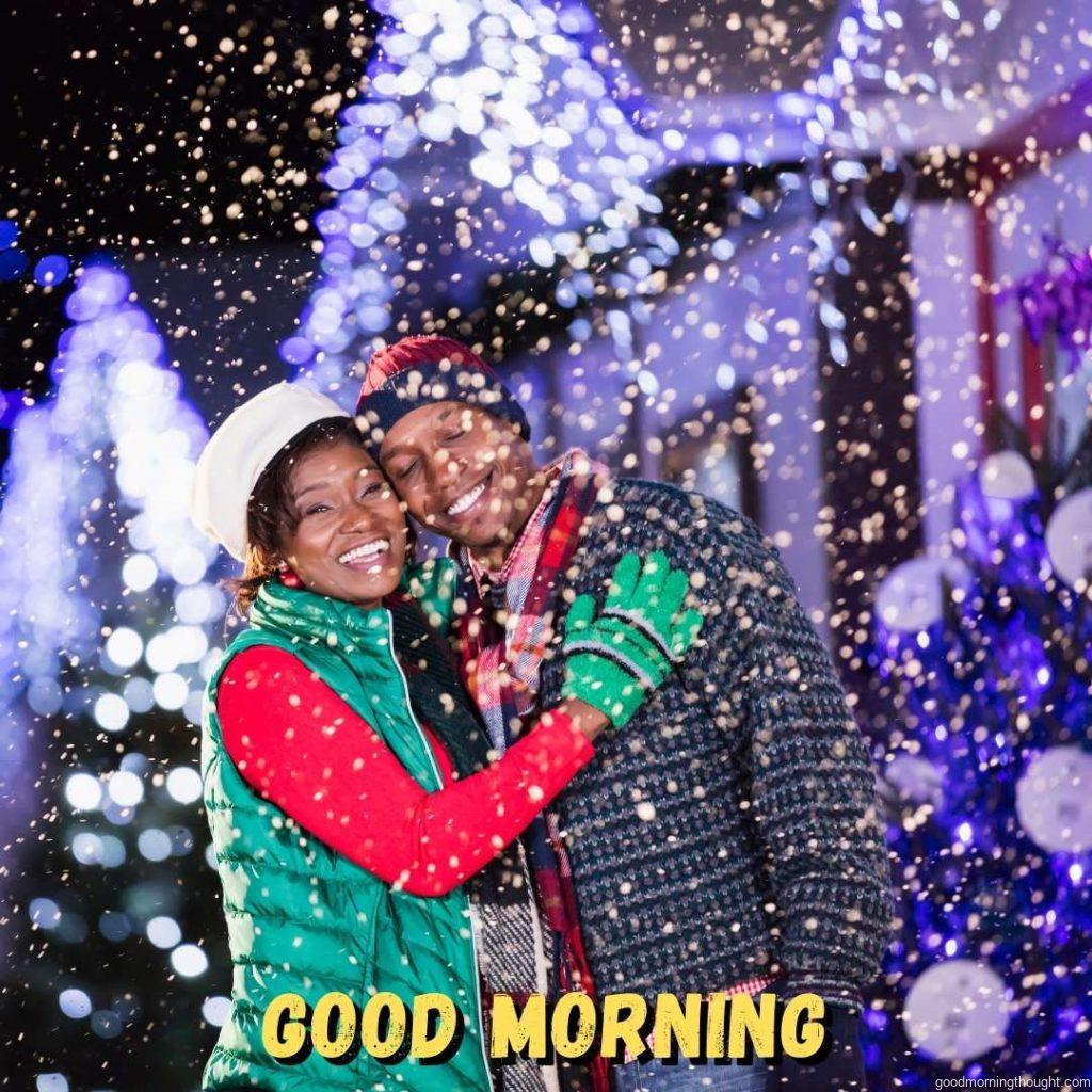 An African-American couple stands outdoors at night, having fun at a winter festival. They are standing in front of buildings decorated with string lights. Good Morning Text