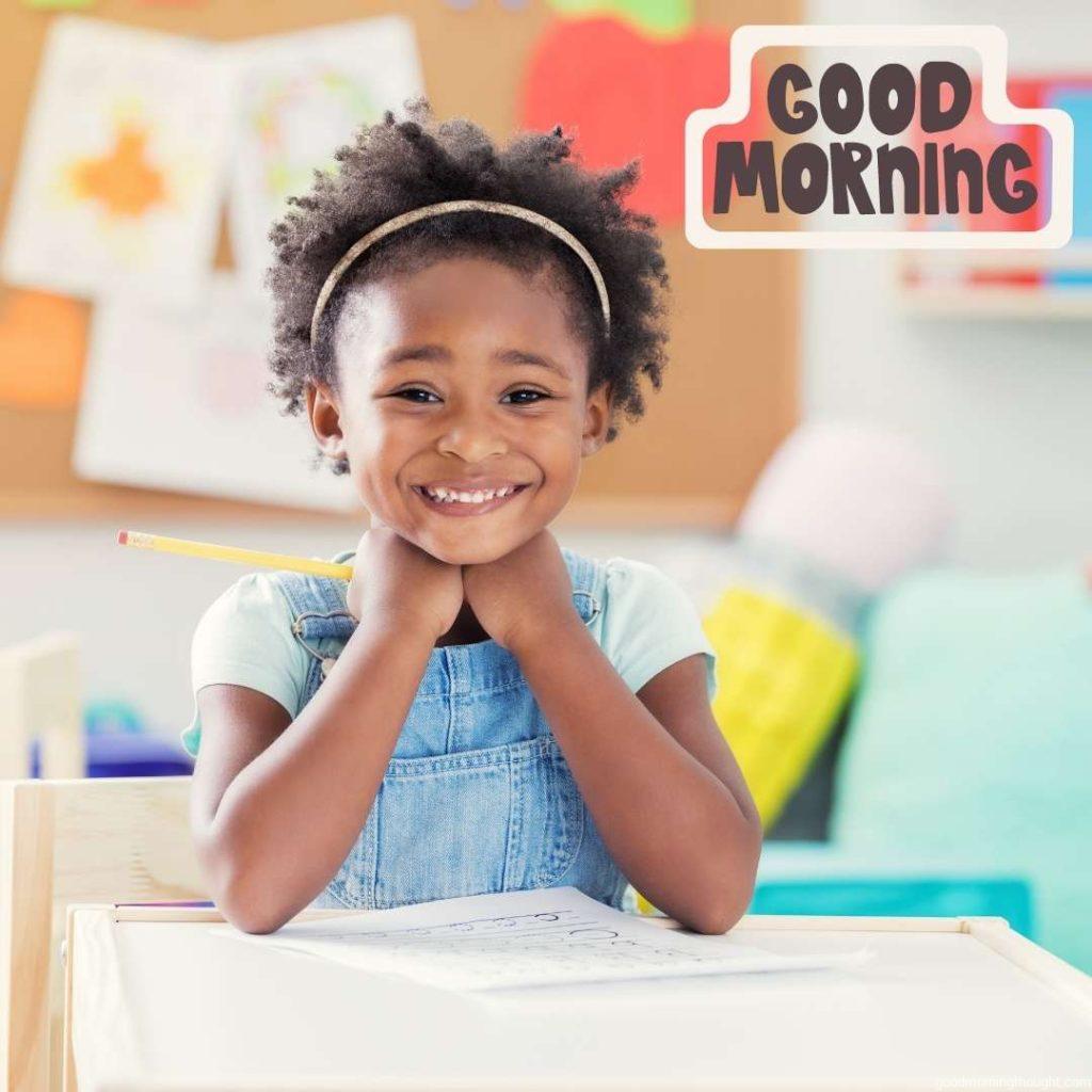 An adorable African American little girl works on a worksheet during preschool. She is smiling at the camera. African American Good Morning Images