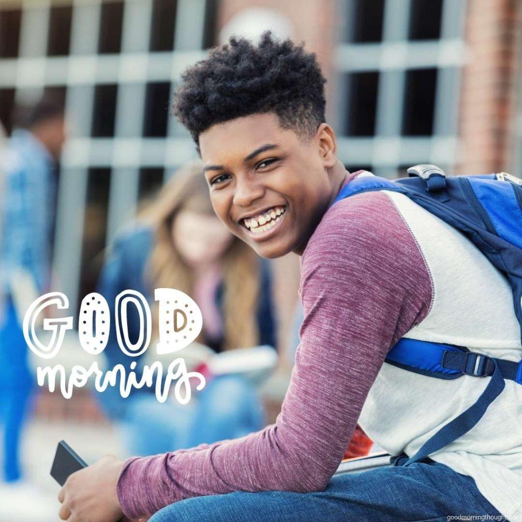 Cheerful African American teenage boy waits outdoors on the school campus. He is holding a cell phone. Good Morning Text