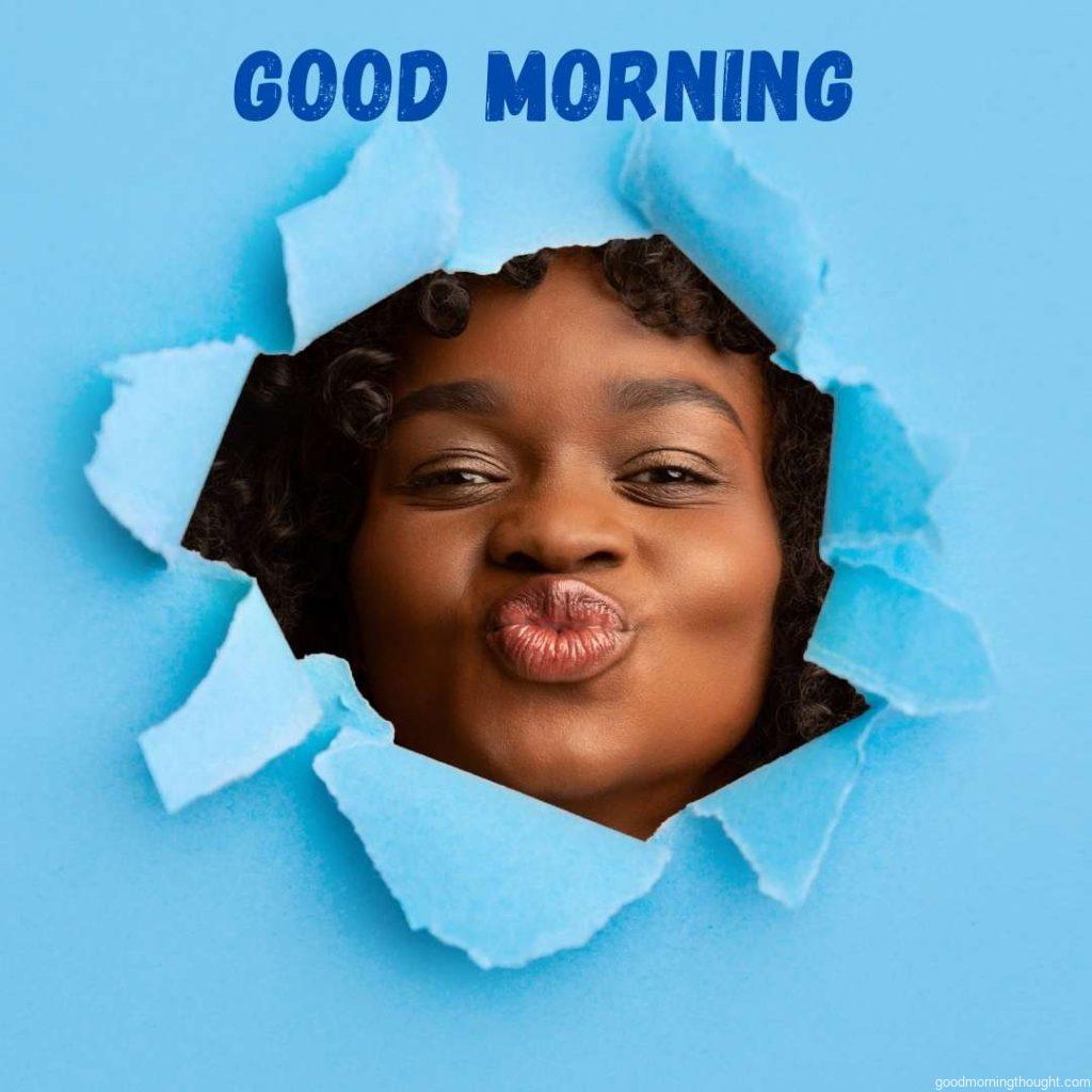 Funny, pretty happy millennial African American woman blows a kiss and looks through a hole in blue paper. Facial expression, close up, Good morning text