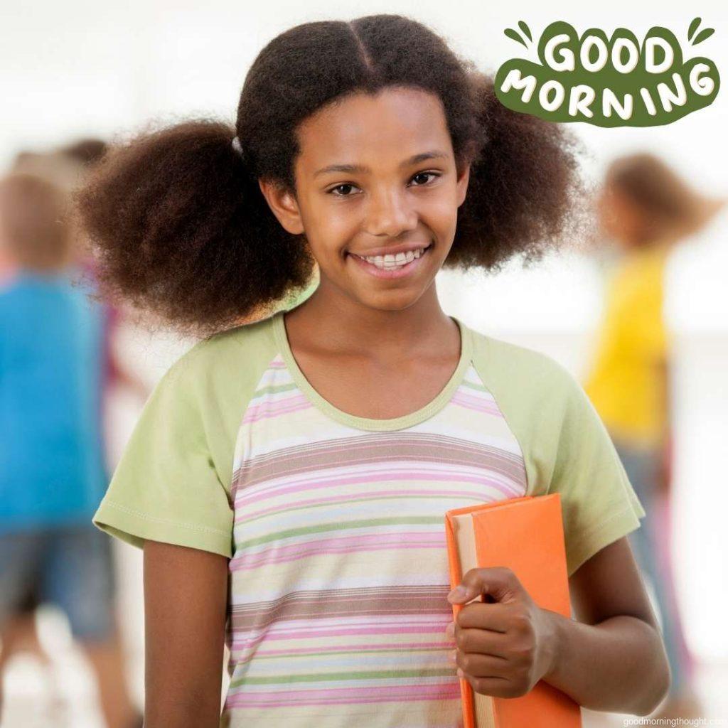 Portrait of a smiling African American schoolgirl holding a book and looking at the camera Other children are in the background. Good Morning Text